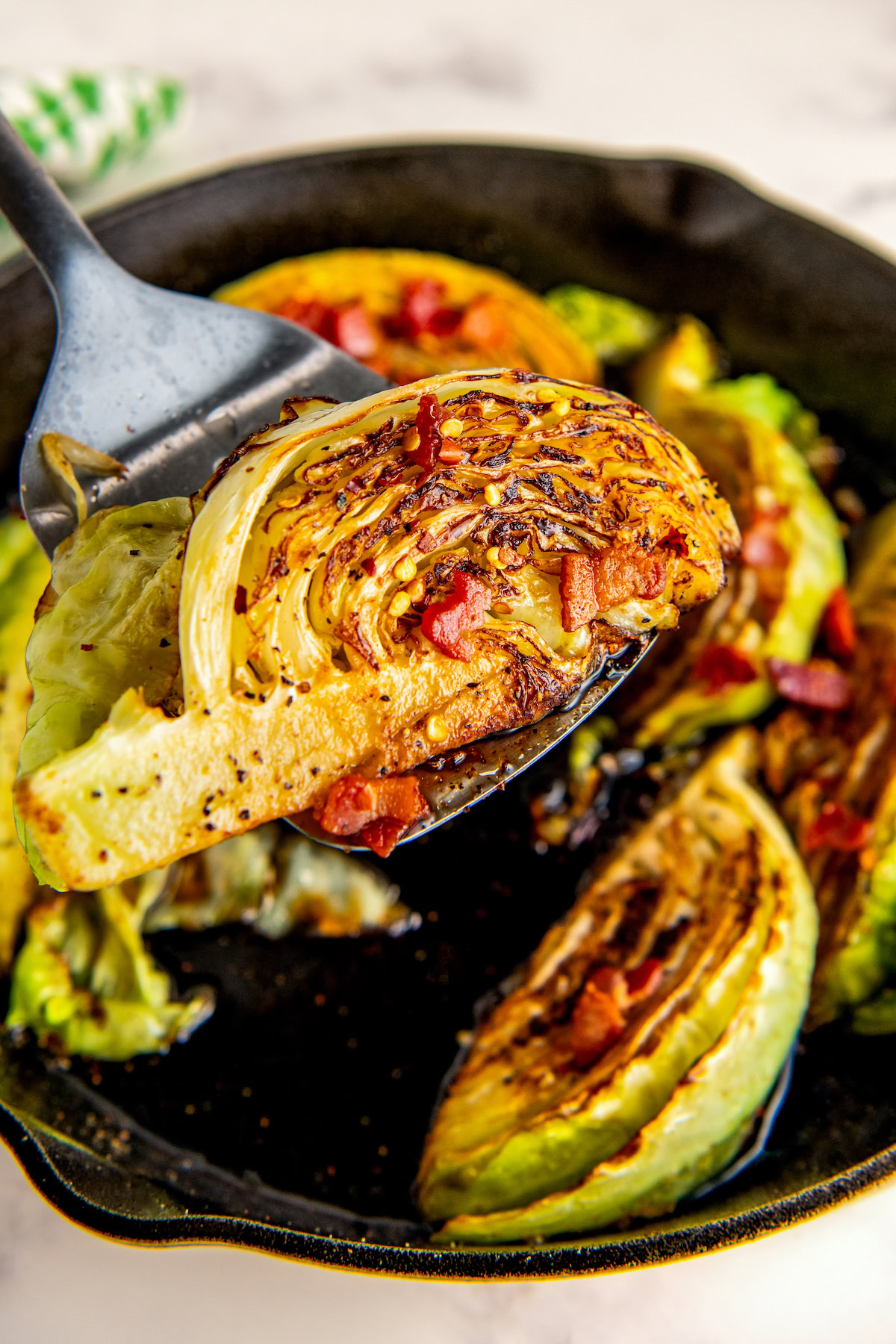 Sliced fried cabbage with crispy bacon and hot honey being scooped out of a cast iron skillet with a spatula. 