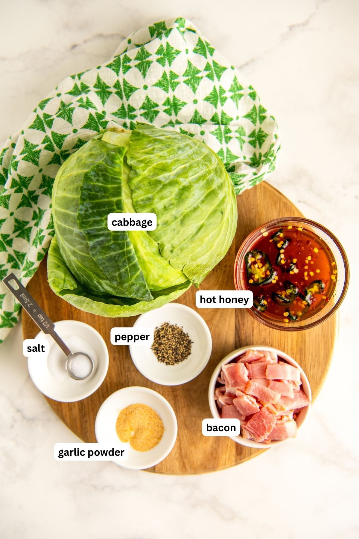 Ingredients for fried cabbage recipe arranged on a cutting board. From top to bottom: green cabbage, hot honey, salt, pepper, bacon and garlic powder. 