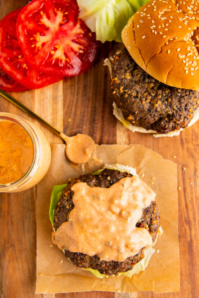 A partially assembled hamburger topped with In and Out burger sauce, next to tomato slices on a wooden cutting board.