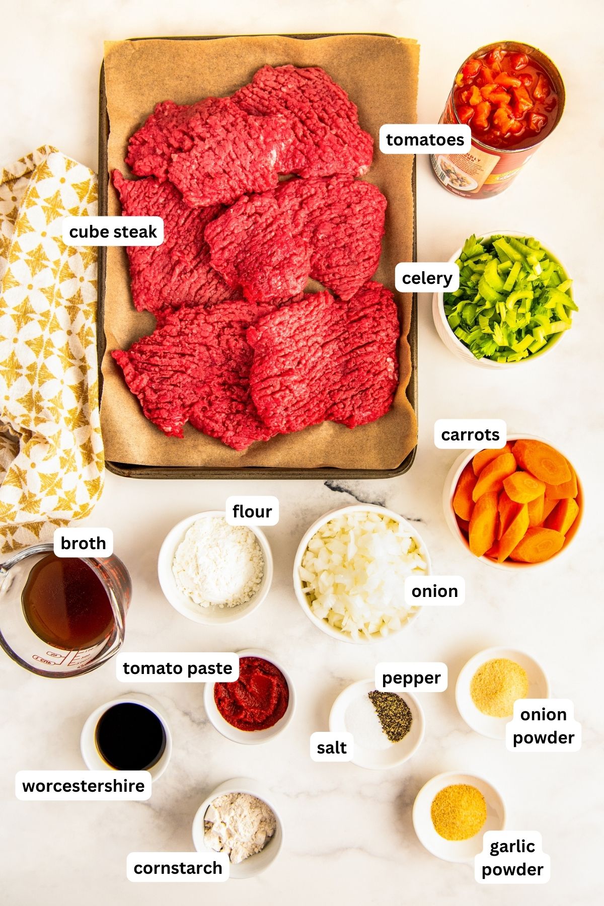 Labeled ingredients for crockpot Swiss steak recipe in bowls. From top to bottom: canned diced tomatoes, cube steak, celery, carrots, onion, flour, broth, tomato paste, salt, pepper, onion powder, Worcestershire sauce, cornstarch and garlic powder. 