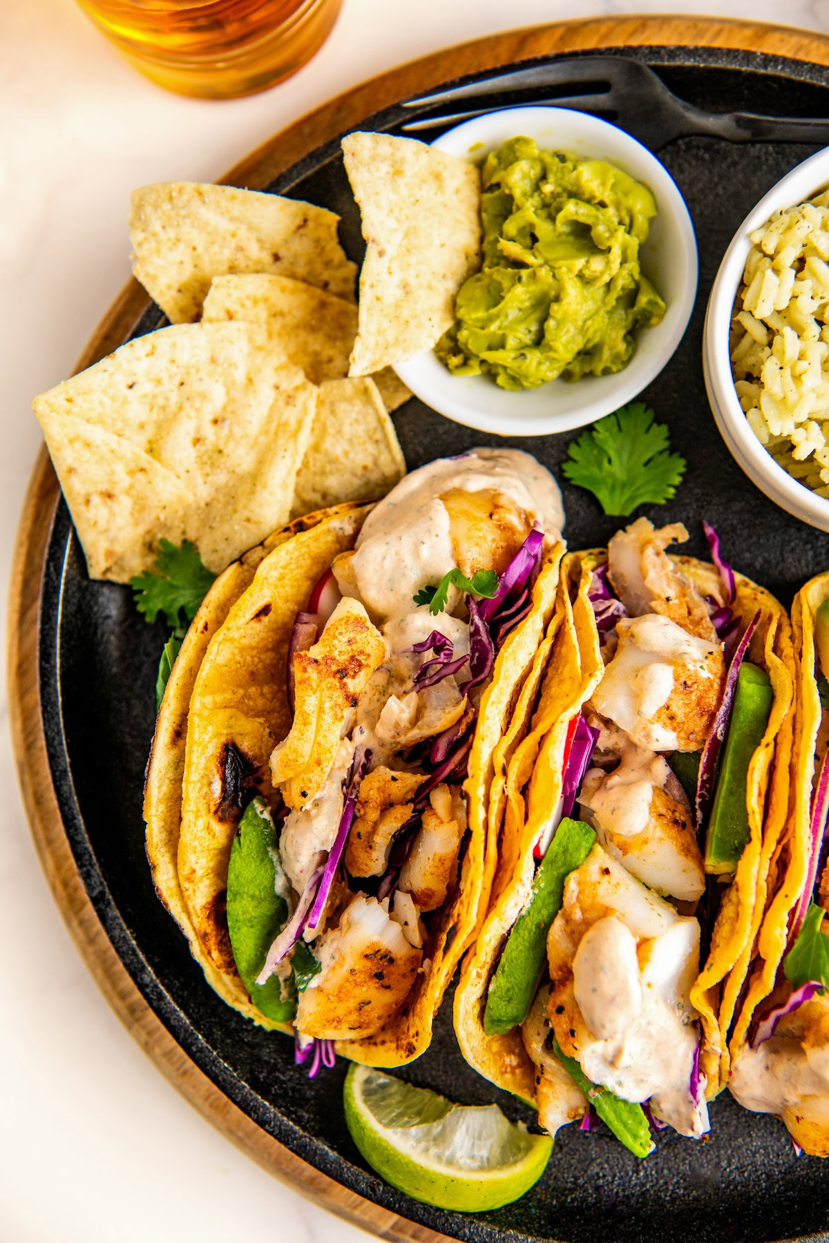 Creamy fish taco sauce drizzled over fish tacos on a plate with chips and guacamole on the side.