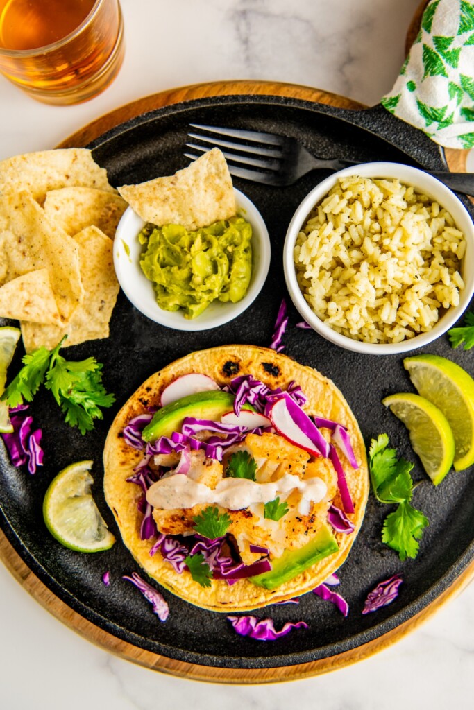 A corn tortilla taco with fish on a plate being assembled and topped with fresh avocado, purple cabbage, radishes and a creamy chipotle sauce.