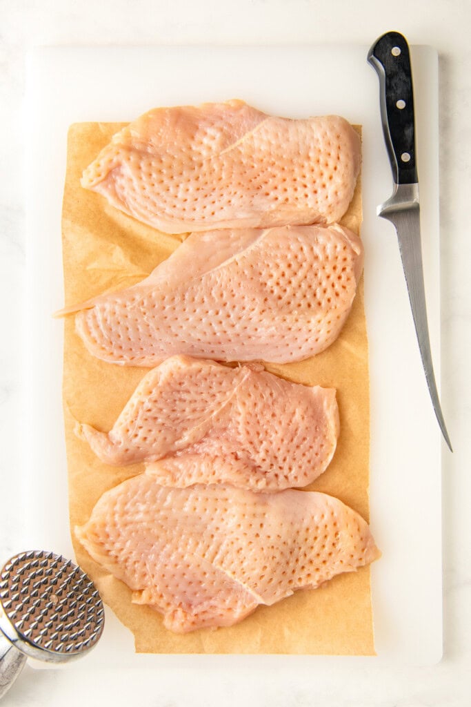 Tenderized chicken breasts on a cutting board with a meat mallet on the side.