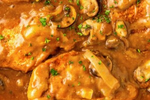 A serving spoon scooping out a tender and juicy smothered pork chop in gravy with mushrooms out of a skillet.