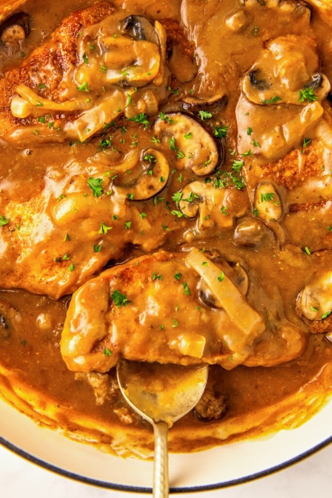 A serving spoon scooping out a tender and juicy smothered pork chop in gravy with mushrooms out of a skillet.