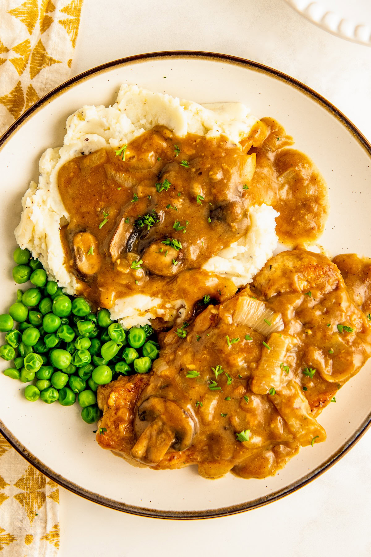 Smothered pork chops and gravy served on a plate with a side of mashed potatoes and peas.