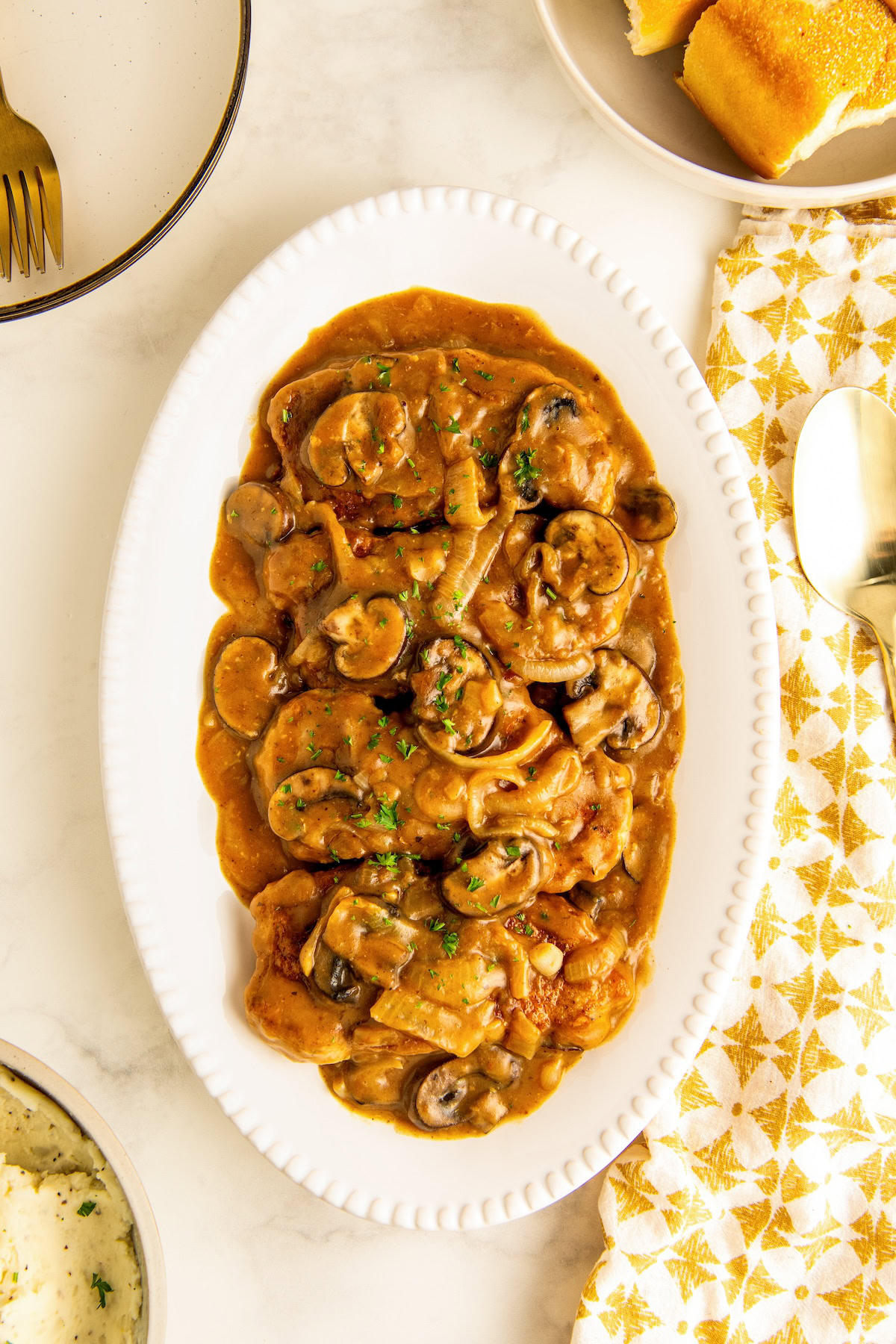 A serving plate filled with tender smothered pork chops with mushroom gravy topped with chopped fresh herbs.