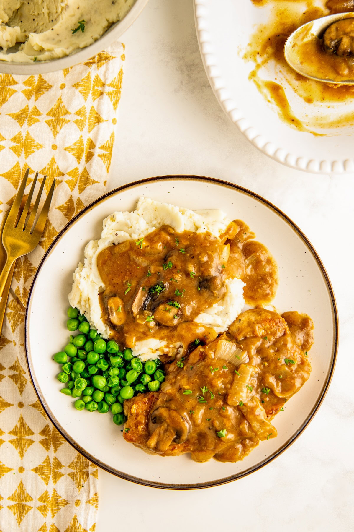 Tender and juicy smothered pork chops in a rich, brown gravy with mushrooms and onions served over mashed potatoes with a side of green peas.