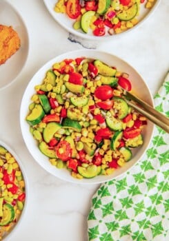 A bowl of zucchini succotash with cherry tomatoes, corn and lima beans with two spoons on the side for serving.
