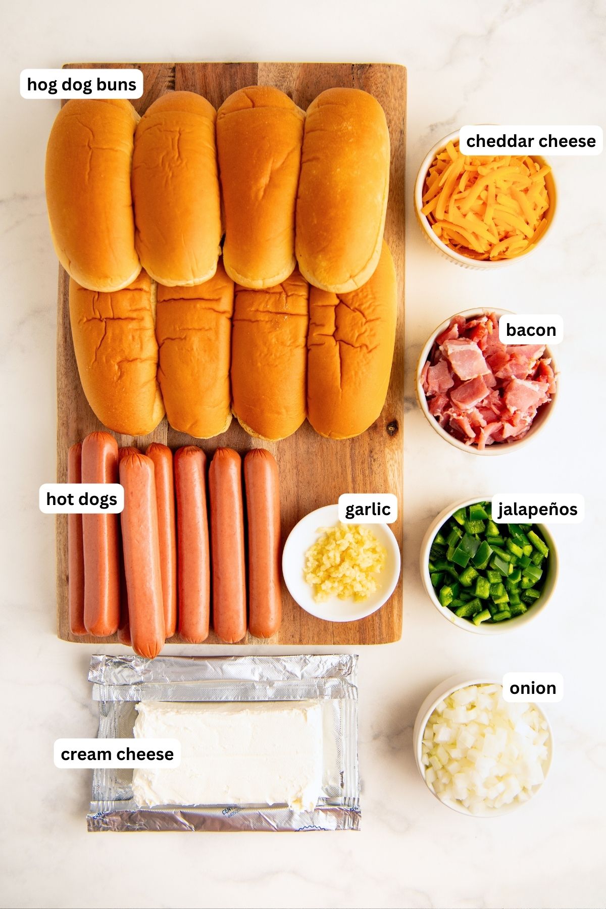 Ingredients for jalapeno popper hot dog recipe arranged in bowls and on a cutting board. From top to bottom: hot dog buns, cheddar cheese, bacon, hot dogs, garlic, jalapeño, creamy cheese and onion.