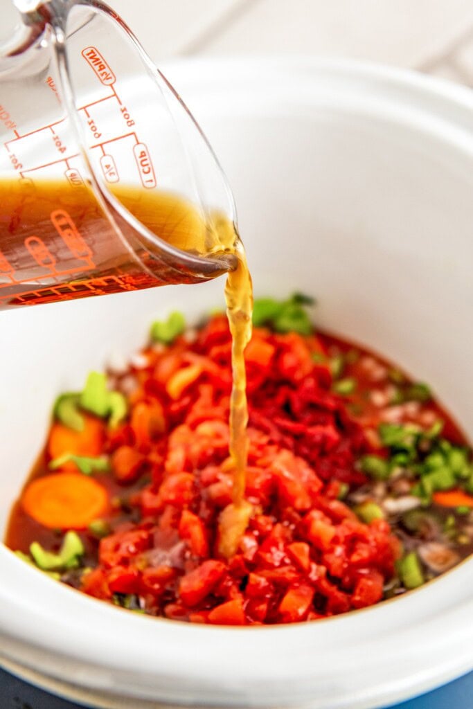 Beef broth being poured into the crockpot filled with Swiss steaks and vegetables.