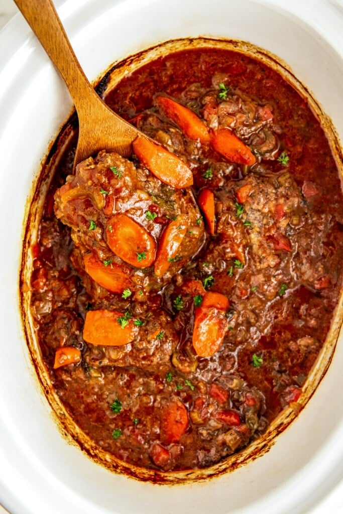 A wooden spoon scooping up a cooked Swiss steak from crockpot.