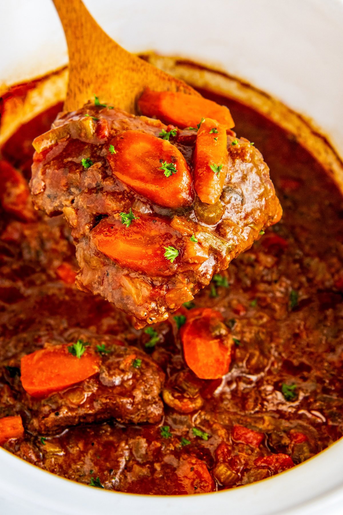 A wooden spoon lifting a serving of tender crockpot Swiss steak from a slow cooker.