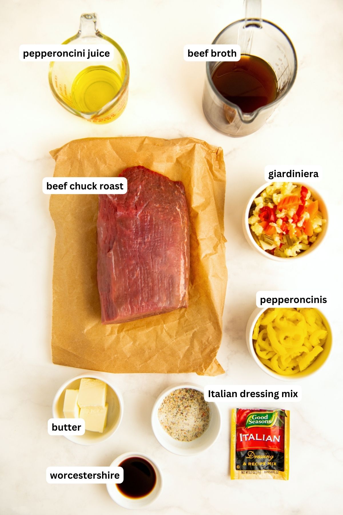 Labeled ingredients for crock pot Italian beef sandwiches recipe. From top to bottom: pepperoncini juice, beef broth, beef chuck roast, giardiniera, pepperoncinis, butter, Italian dressing mix and worcestershire. 