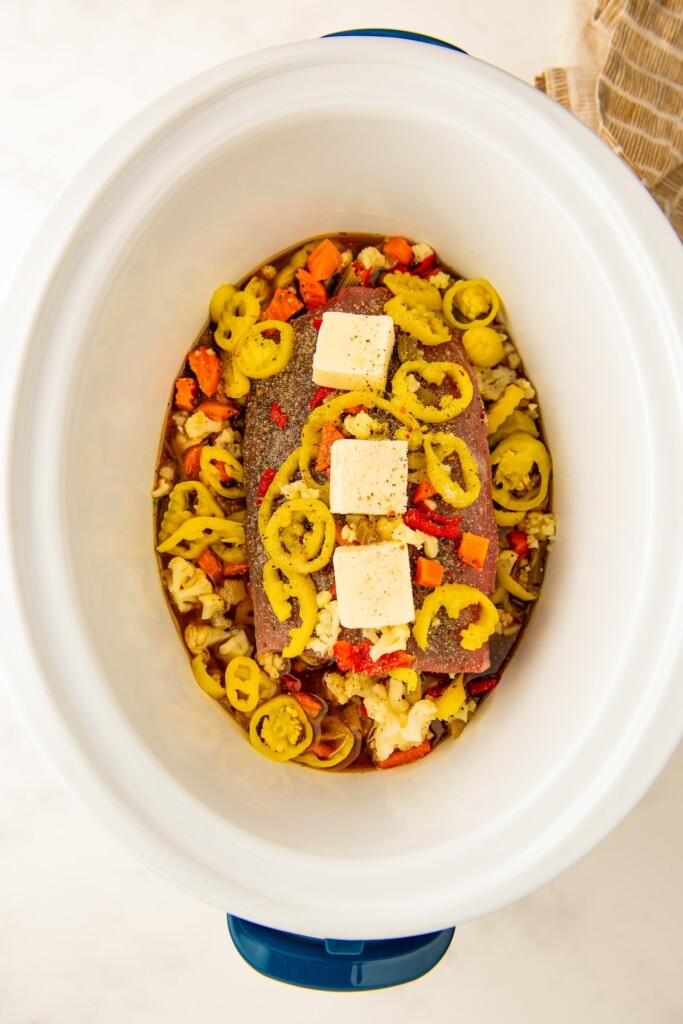 A chuck roast is in a Crockpot with butter, peppers, seasonings and broth. 