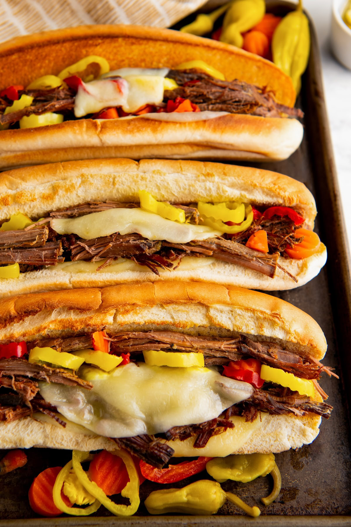 A close-up of three slow cooker Italian beef sandwiches in toasted hoagies with melted provolone and giardiniera veggies. 