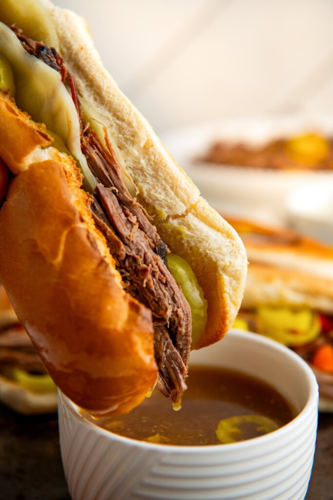 An Italian beef sandwich being dipped into a bowl of warm and savory au jus. 