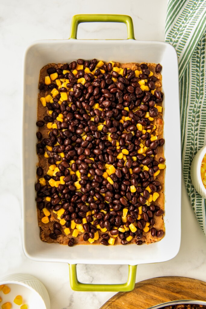 Black beans and corn being layered on top of refried beans in a casserole dish.