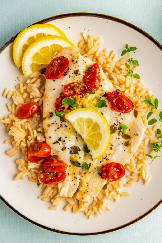 Plate of flaky fish with juicy tomatoes and lemon slices over fluffy rice.