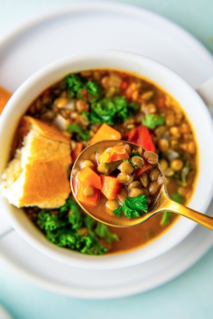 A bowl of tomatoey lentil soup with kale and fresh parsley on top with a spoon lifting a bite of soup.