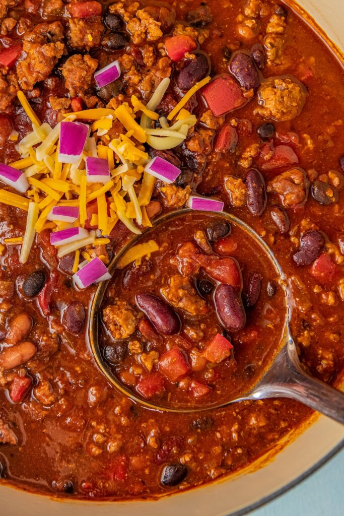 A large pot of ground turkey chili with three kinds of beans with a ladle scooping out a serving.