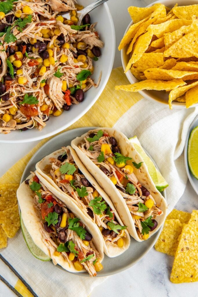 Shredded mexican chicken served in tortillas with fresh cilantro with a bowl of crockpot mexican chicken in the background.