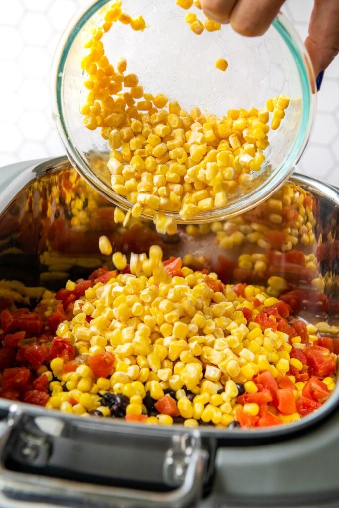 Corn being poured into a crockpot filled with chicken, taco seasoning, black beans and rotel.