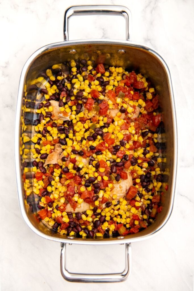 A slow cooker filled with chicken breasts, taco seasoning, black beans, corn and rotel.