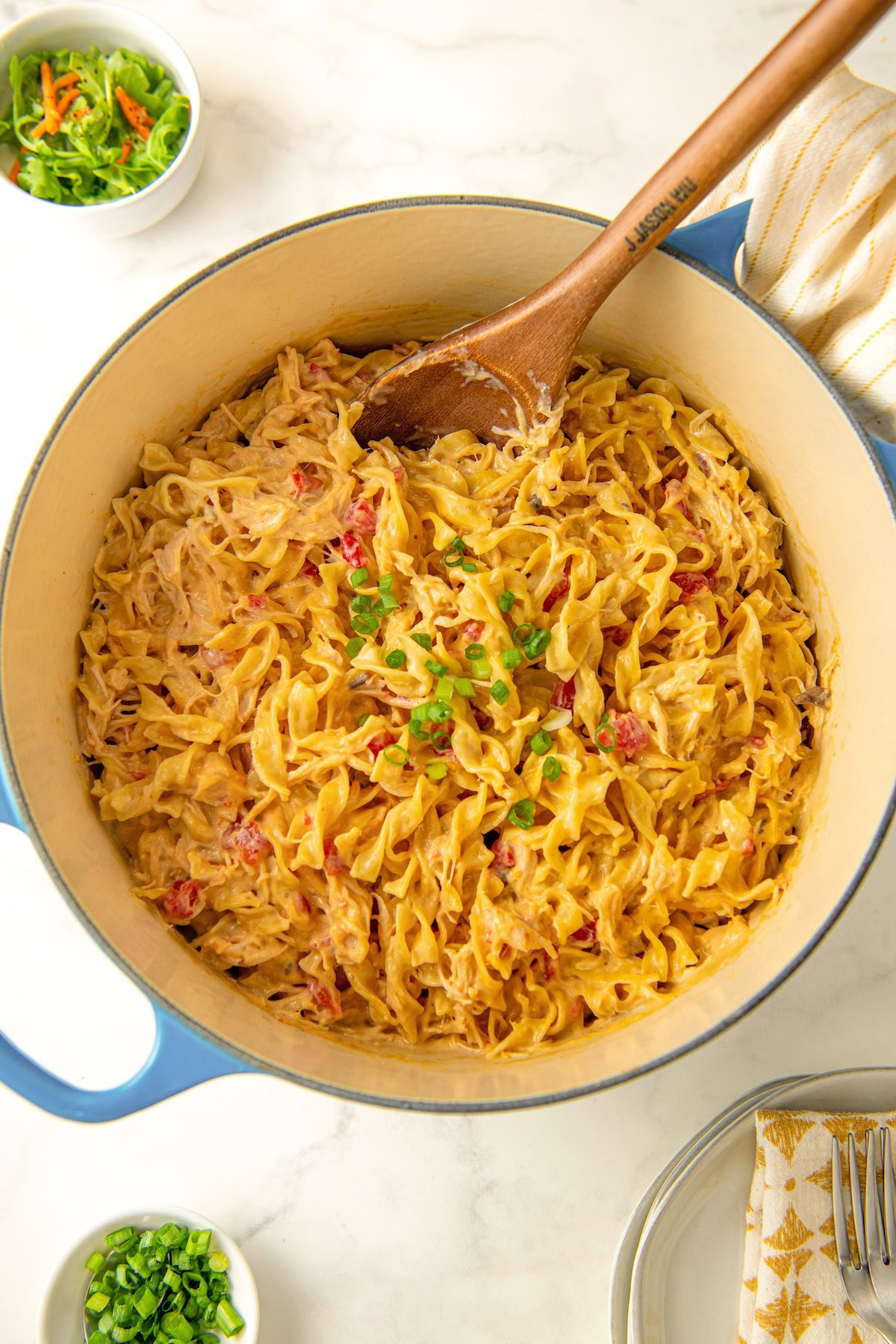 A pot of creamy rotel chicken pasta with egg noodles and green onions on top being mixed with a wooden spoon.