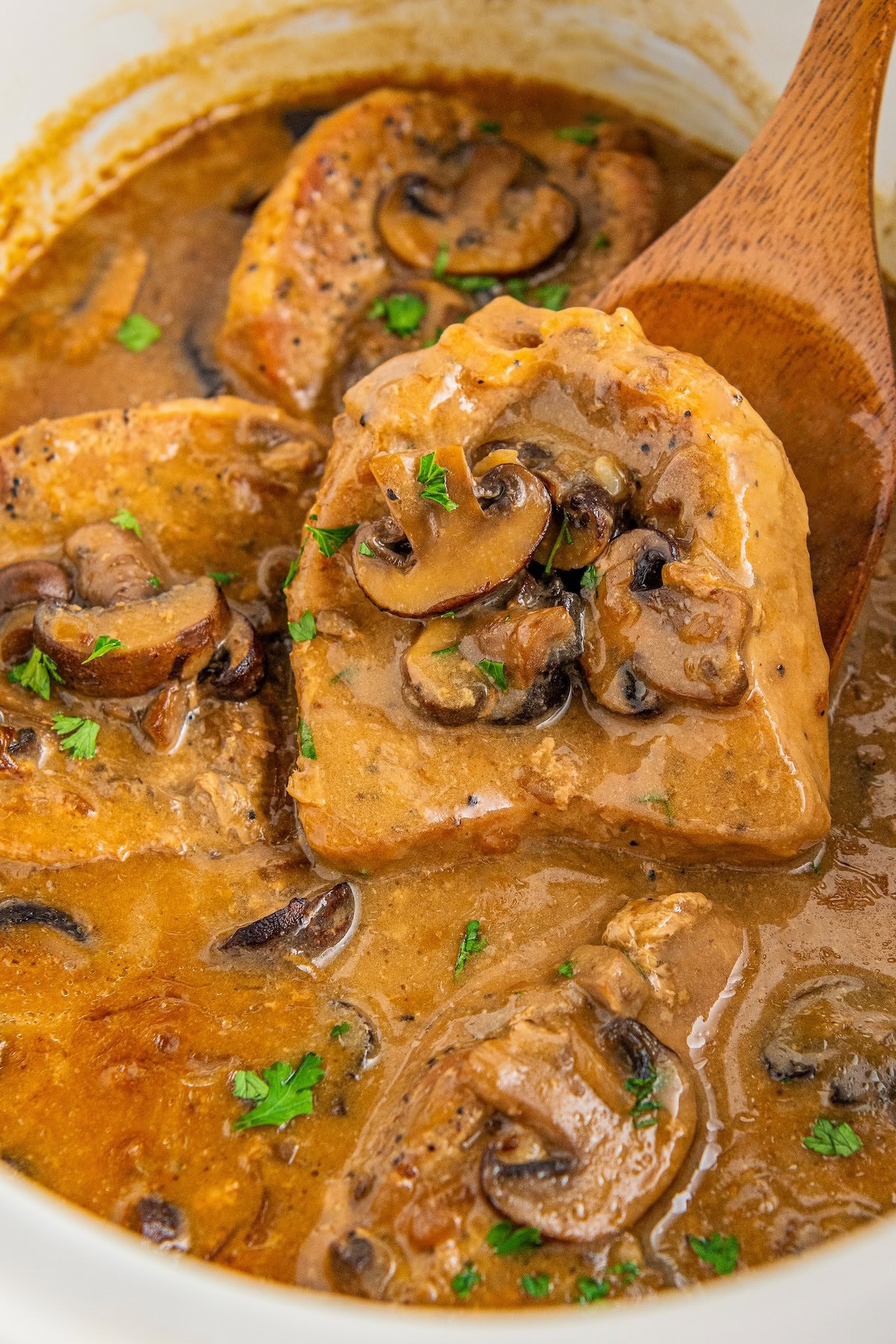 Crock pot pork chops and gravy with mushrooms in a slow cooker with a wooden spoon lifting one of the smothered pork chops.