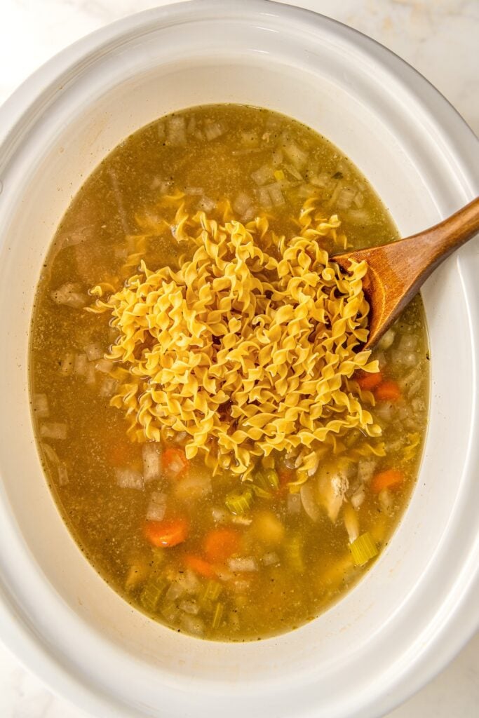 Egg noodles are being added to a crockpot filled with soup.