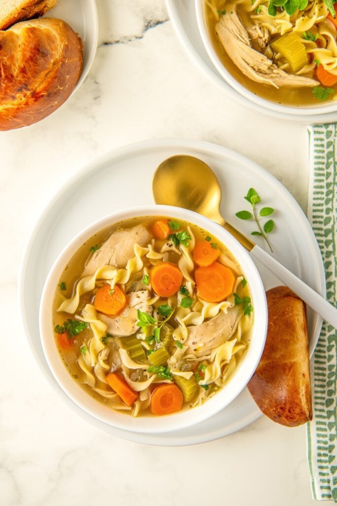 A spoon is placed next to a bowl of slow cooker chicken noodle soup with egg noodles and a hunk of bread on the side.