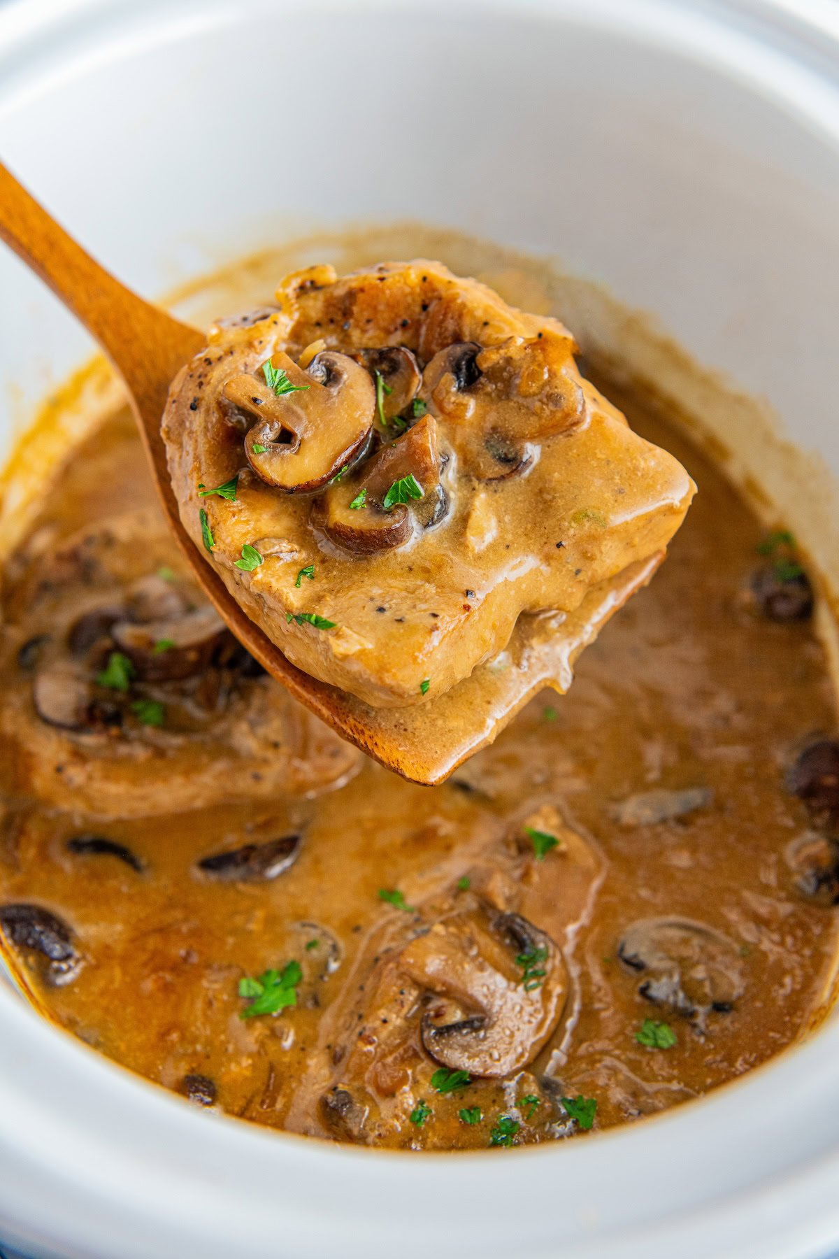 Tender crock pot pork chops with mushroom gravy are being lifted out of a slow cooker with a wooden spoon.