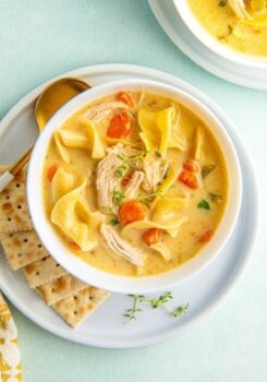 A bowl of rich and creamy chicken noodle soup with egg noodles with crackers and a spoon on the side.