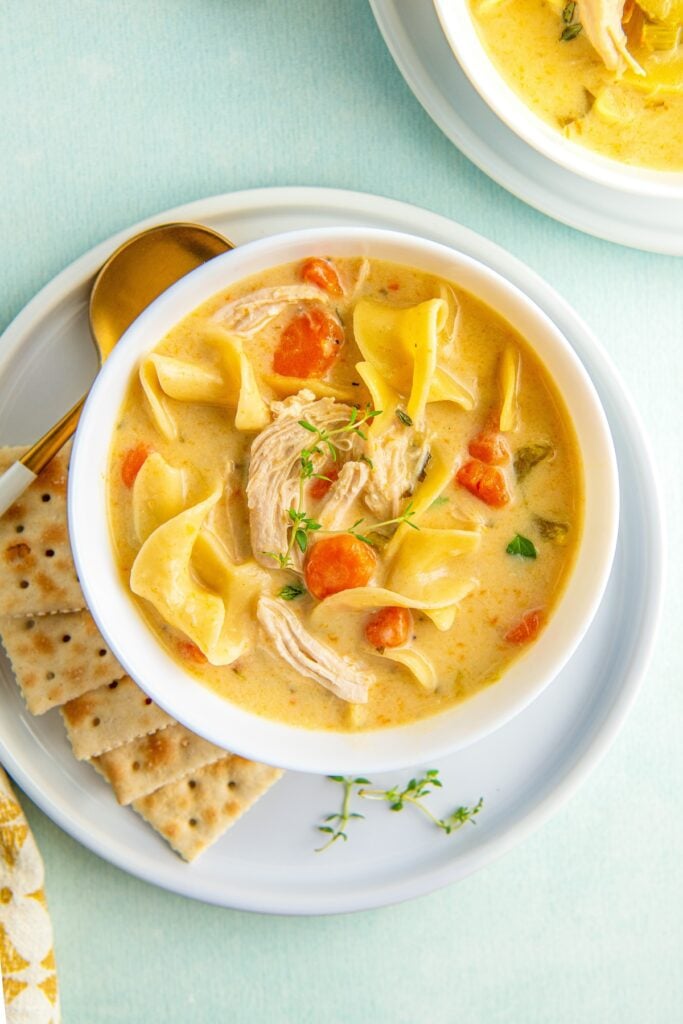 A bowl of rich and creamy chicken noodle soup with egg noodles with crackers and a spoon on the side.