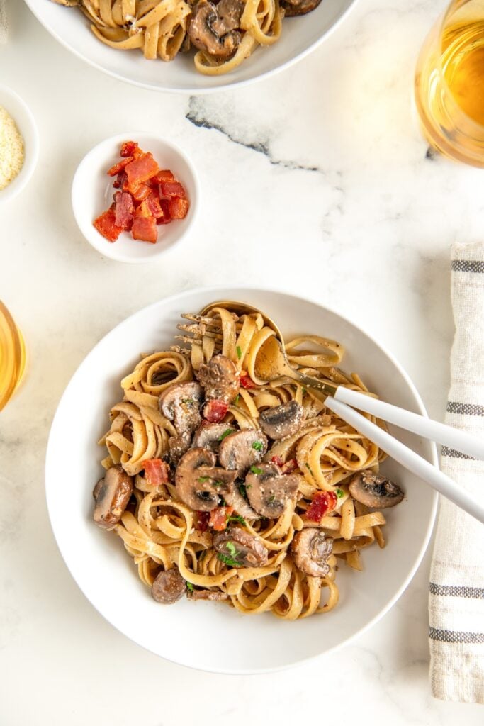 A bowl of creamy pasta with mushrooms next to a small bowl of crispy bacon.