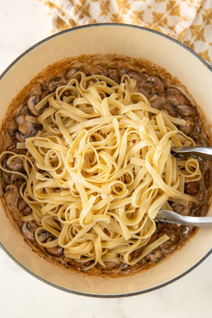 Cooked fettuccini noodles being added to a large pot of cooked mushrooms in a creamy sauce.