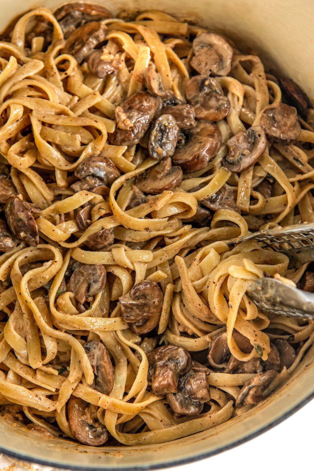 Cooked fettuccini noodles being tossed in a mushroom pasta sauce with tongs.