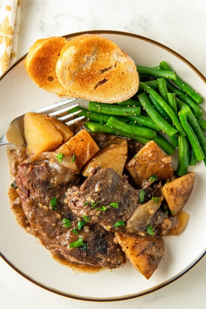 A white plate is topped with green beans, bread, and crockpot steak and potatoes.