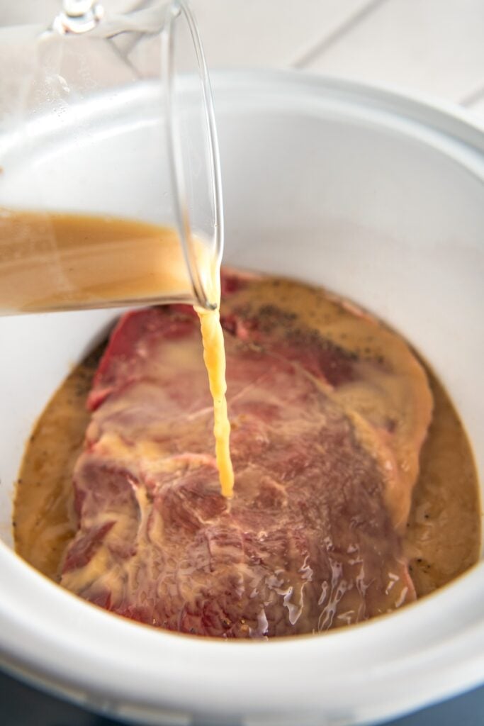 Beef broth is being added to a crockpot filled with steak.