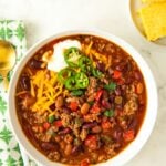 A bowl of savory, hearty beef chili is topped with shredded cheese, sliced jalapeños and sour cream.