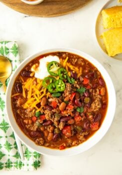 A bowl of savory, hearty beef chili is topped with shredded cheese, sliced jalapeños and sour cream.