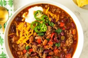 A bowl of savory, hearty beef chili is topped with shredded cheese, sliced jalapeños and sour cream.