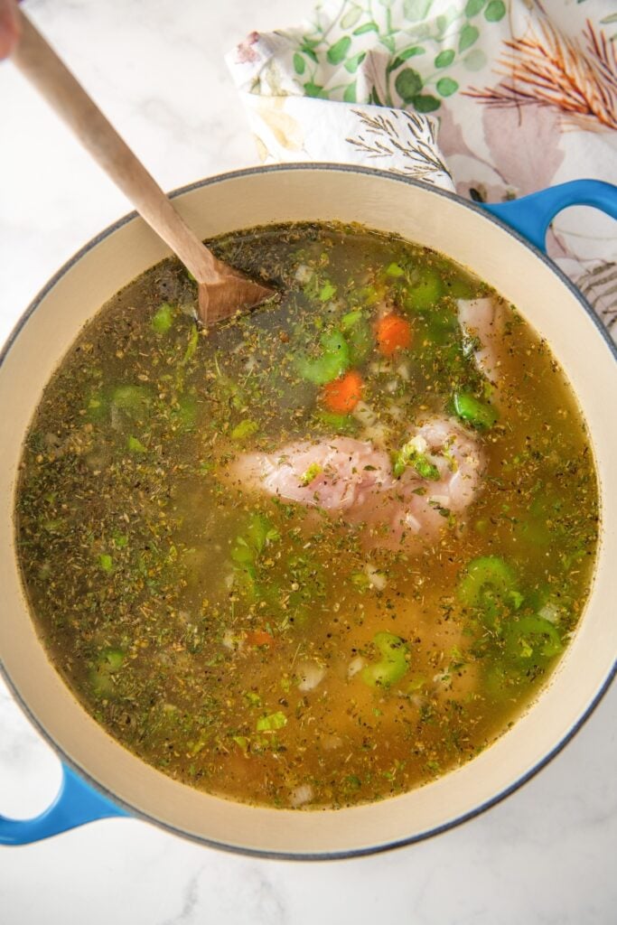 A wooden spoon is stirring chicken lemon rice soup ingredients in a large dutch oven.