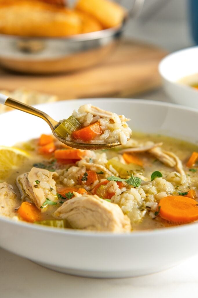 A spoon is lifting a bite sized portion of lemon and chicken soup from a white bowl.