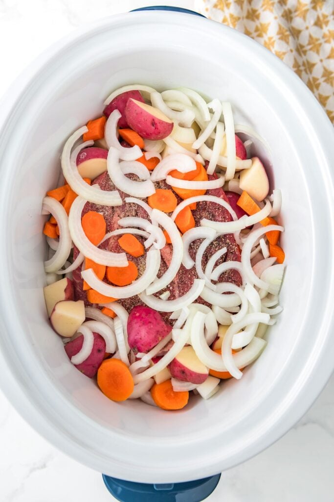 Uncooked vegetables and meat in a white crockpot.