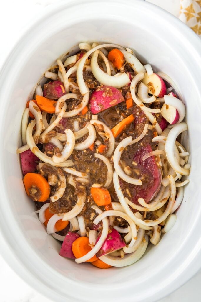 Vegetables, pot roast and seasoned broth are placed in a large white crockpot.