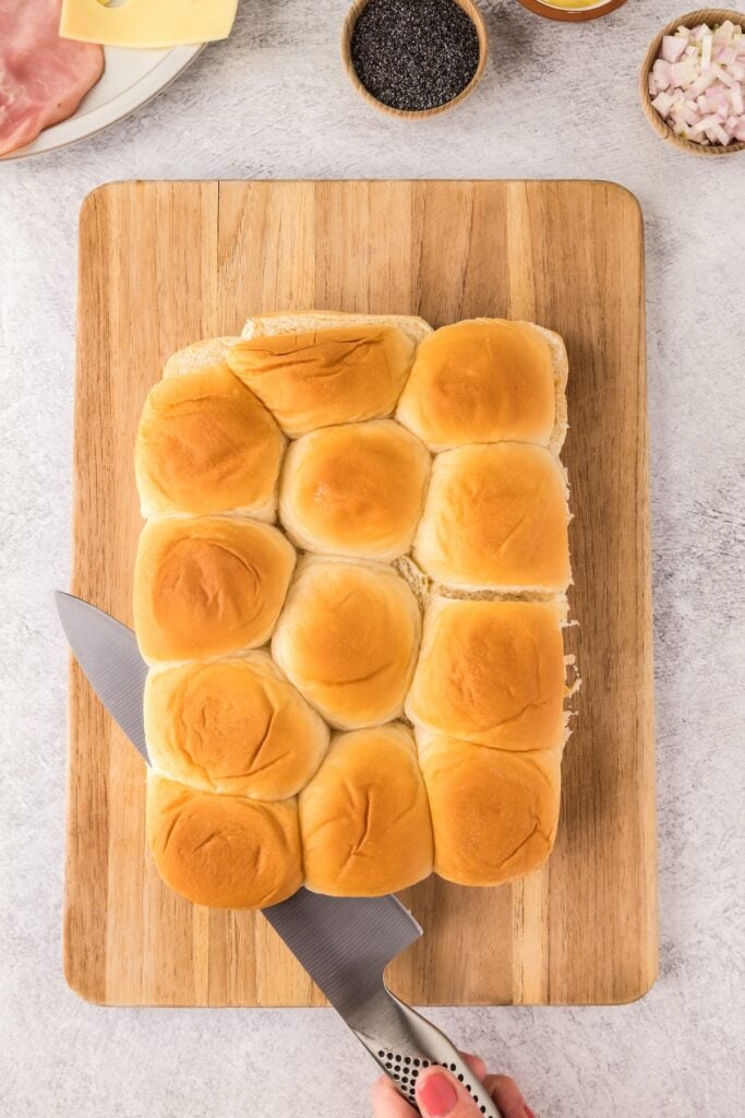 A batch of Hawaiian rolls are being sliced in half with a serrated knife.