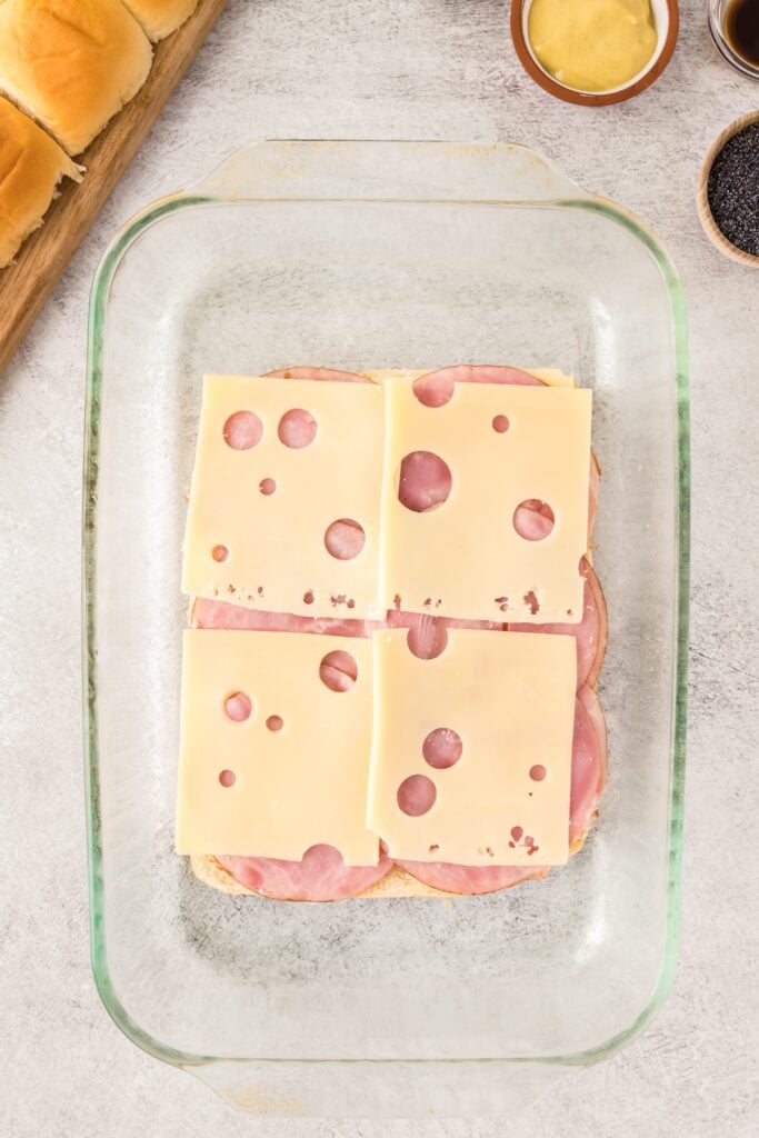 Layers of deli ham and sliced Swiss cheese are layered on top of bread in a baking dish.