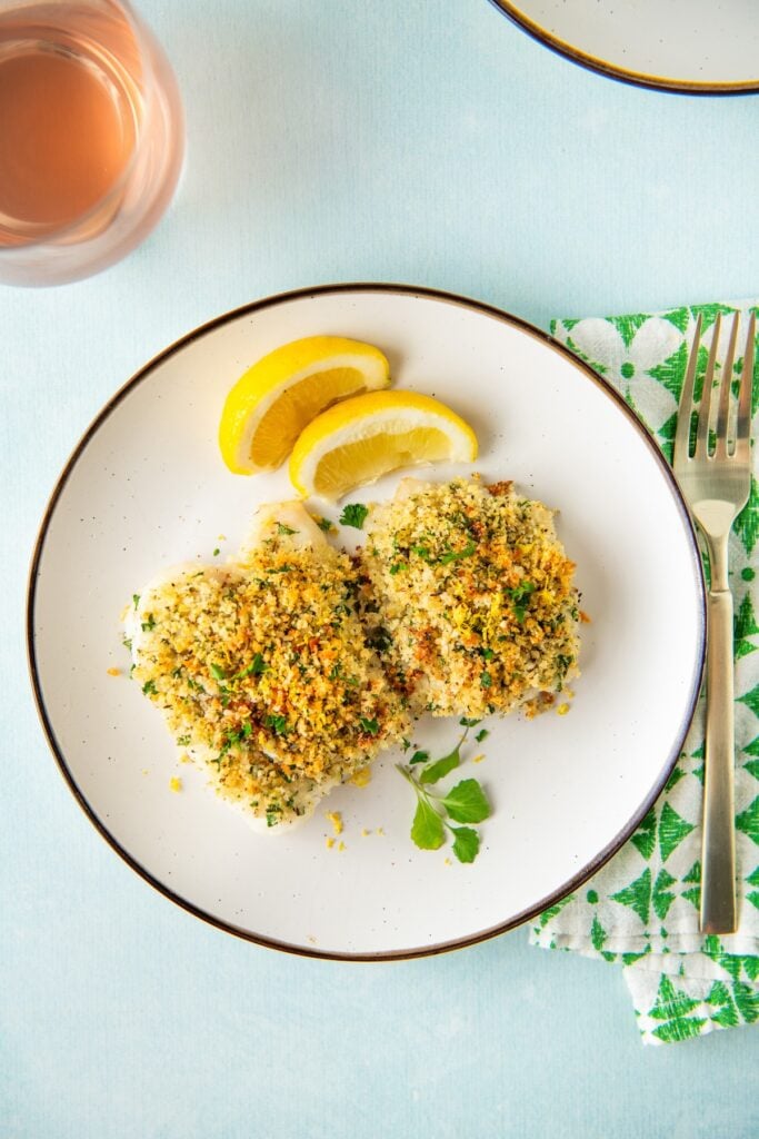 Flaky oven baked cod topped with parmesan and breadcrumbs is plated with lemon wedges.